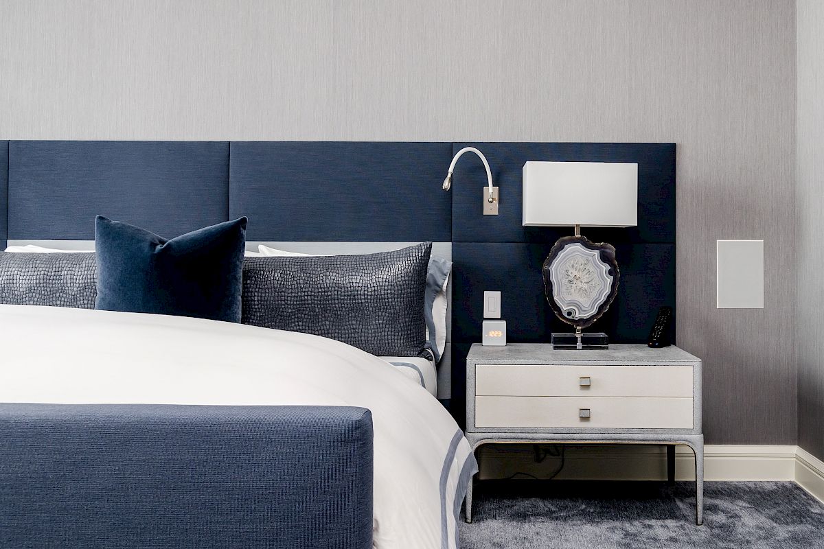 A modern bedroom with a blue upholstered bed, pillows, a bedside nightstand, lamp, wall outlets, and a remote.