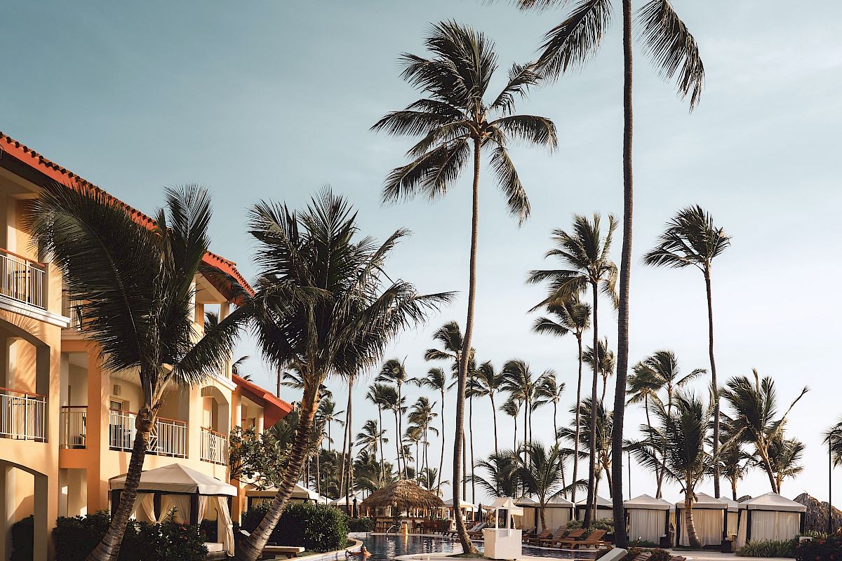 A tropical resort scene with a swimming pool, palm trees, and sun loungers beside hotel buildings and white cabanas.