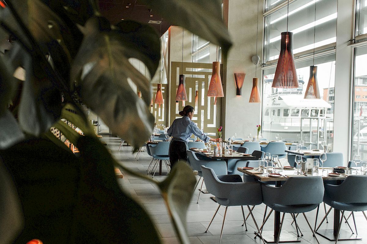 A modern restaurant interior with blue tables set with glassware, warm pendant lights, and a person near the dining area.