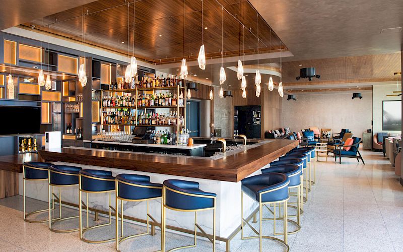 A modern hotel bar with a wooden counter, pendant lights, stocked shelves, and lounge seating.