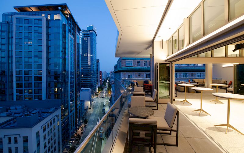 A modern high-rise balcony overlooks a downtown street lined with tall buildings, with patio seating along the edge in early evening light.