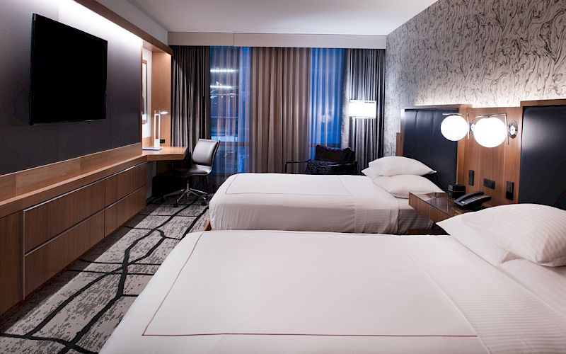 Modern hotel room with two beds, wall TV, desk, and tall window curtains with blue light.