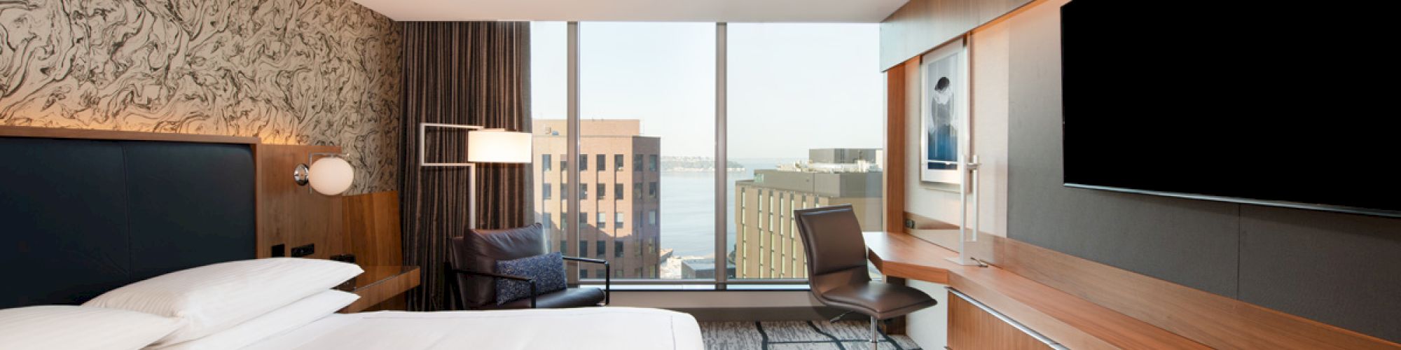 A modern hotel room with a bed, lounge chair, desk setup, flat-screen TV, wood paneling, and large windows overlooking the city.