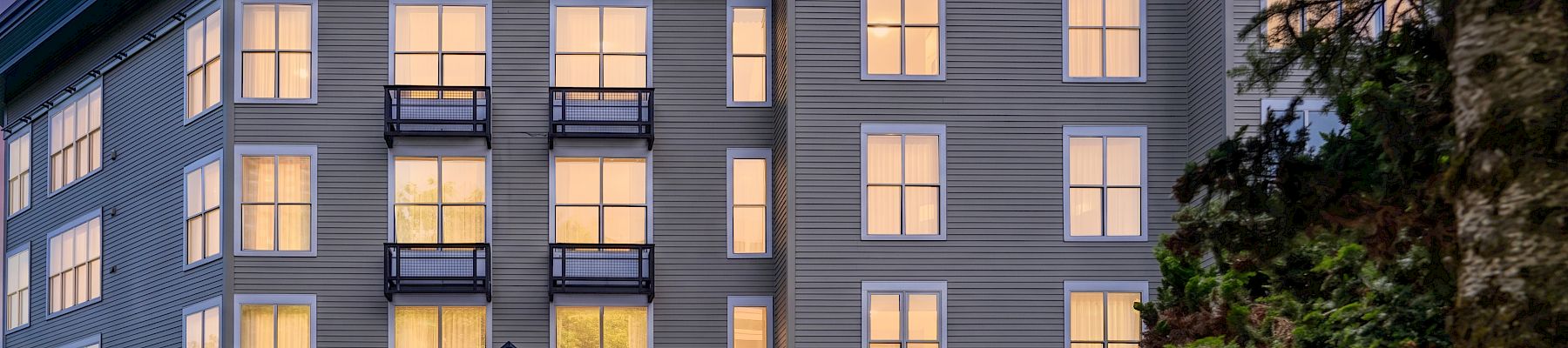 A modern apartment building with lit windows and balconies at dusk, framed by trees and a sign reading &ldquo;RiverPlace&rdquo;.