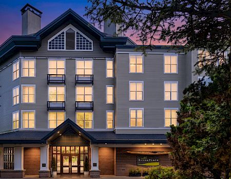 A modern apartment building with lit windows and balconies at dusk, framed by trees and a sign reading “RiverPlace”.